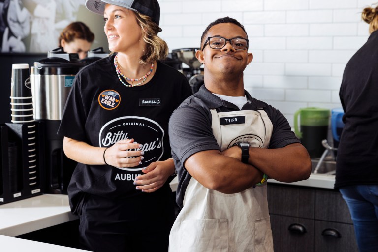 A smiling Bitty & Beau’s Coffee employee wearing an apron, standing behind the counter, representing the mission of inclusion and empowerment through fundraising